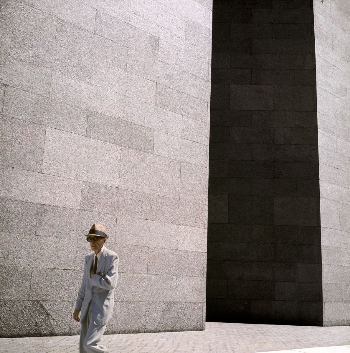 La photographie montre un homme chapeauté marchant devant un immeuble.